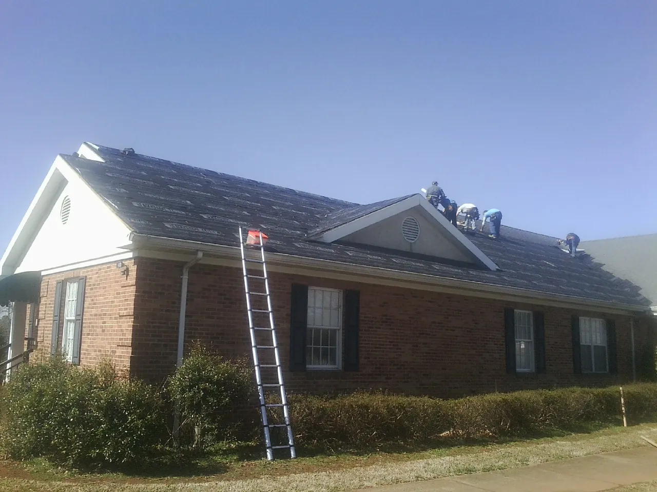 Crew tearing shingles off a brick ranch roof with a ladder set — Emergency Roof Repair work by our crew in Glenham