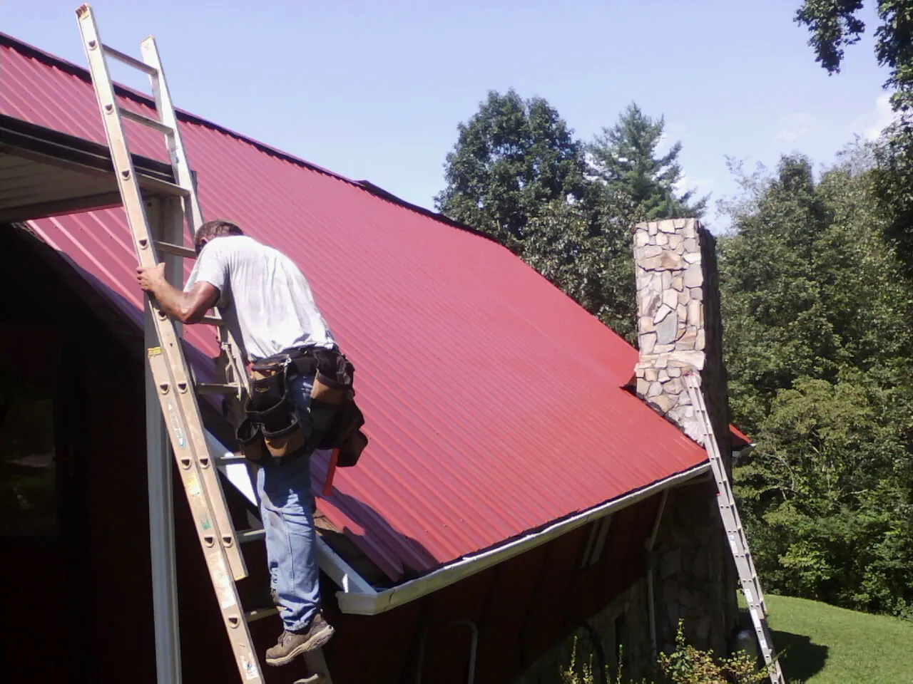 Roofer on a ladder beside a red metal roof and stone chimney — Soffit & Fascia Repair by Roofing Brothers in Glenham